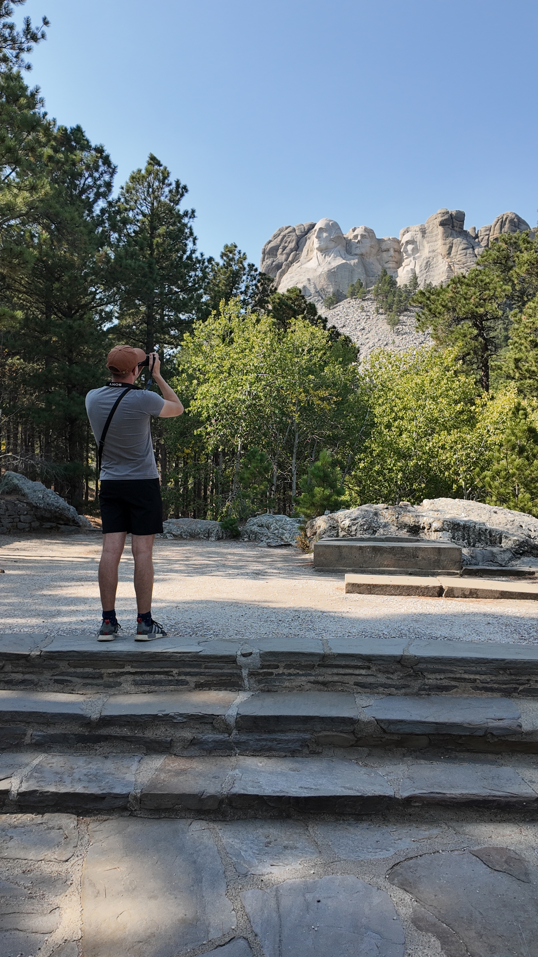 A man taking a photo of Mount Rushmore from the presidential trail. The incredible landscape is surrounded by autumnal trees.